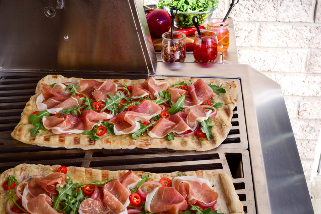 An image of two grilled flatbread with Proscuitto, arugala, red chilis and onions on a grill