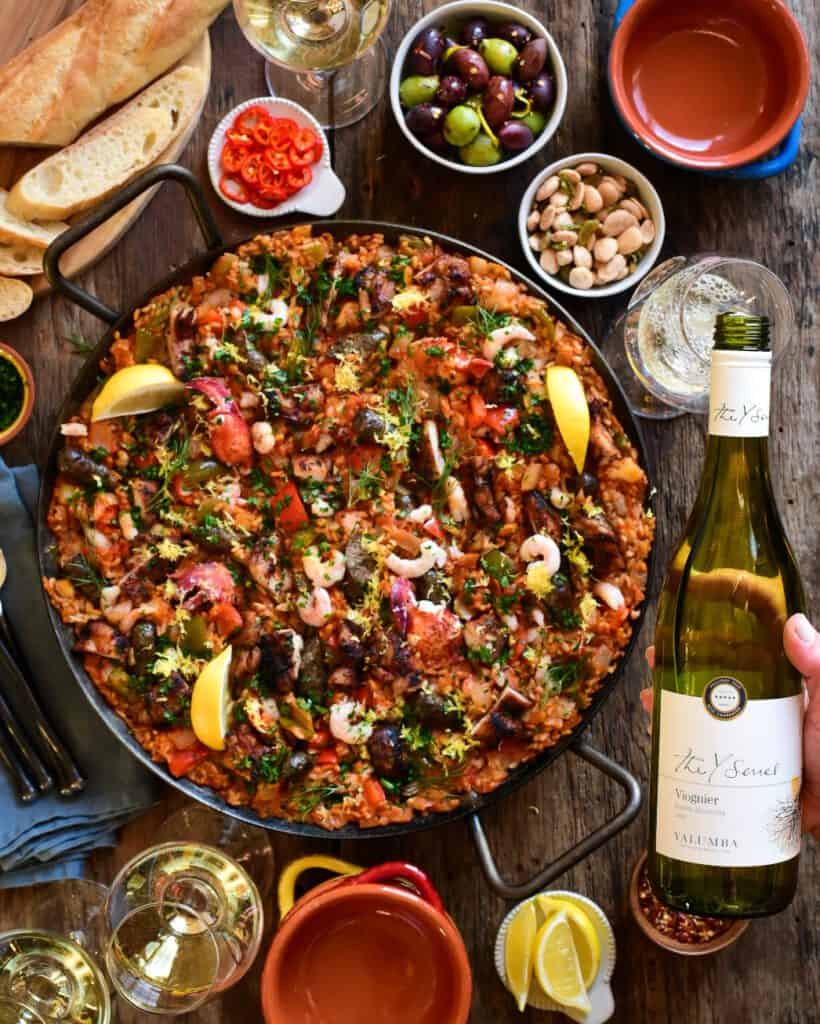 A large, colourful pan of paella filled with seafood, poultry, rice and veggies. This pan is surrounded by a bottle of Viognier, glasses of wine, nuts, olives, lemons and bowls. 