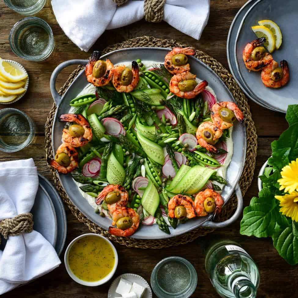 A top down image of a spring salad with grilled wild shrimp, ribbons of cucumber, snap peas, radishes, asparagus tips and green onions . Plates, napkins, wine, glasses, and sauce all surround this dish. 