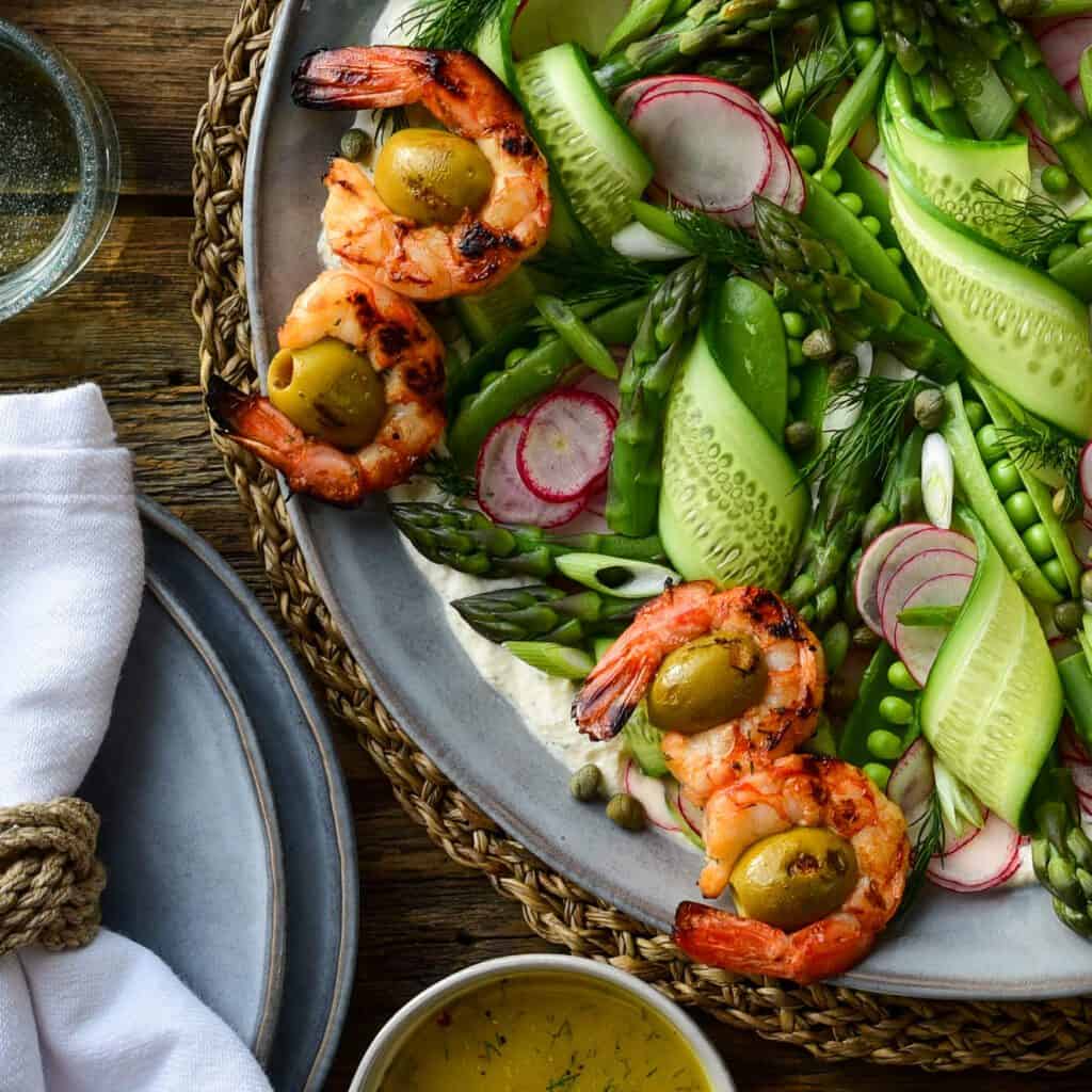 A close up of a top down image of a spring salad with grilled wild shrimp, of ribbons of cucumber, snap peas, radishes, asparagus tips and green onions.