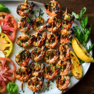 Four sweet and spicy grilled shrimp skewers served with grilled lemons and a tomato salad.