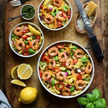 A large and two small bowls of shrimp pasta on a wooden cutting board. Also on this board are sliced lemons, basil, cheese and a grater.