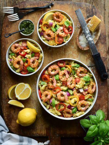 A large and two small bowls of shrimp pasta on a wooden cutting board. Also on this board are sliced lemons, basil, cheese and a grater.