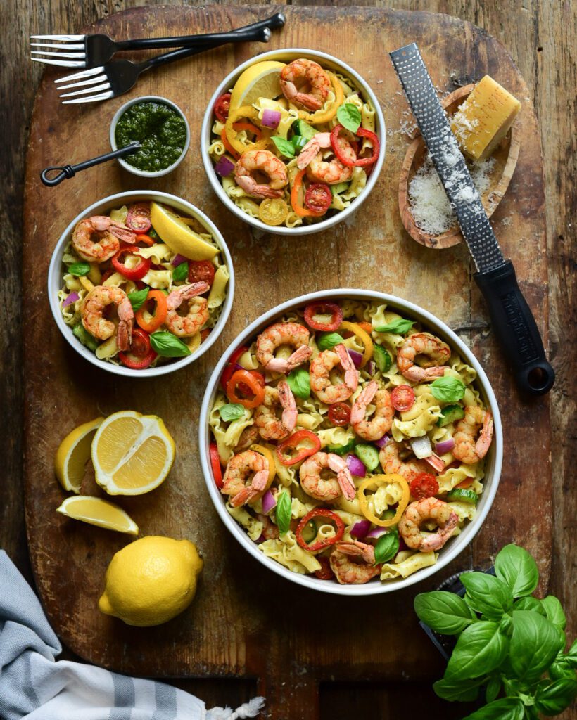 A top down image of a large and two smaller bowls of a Wild Argentinian Shrimp Pesto Pasta Salad on a wooden board. Basil, lemon's and Parmesan cheese. 