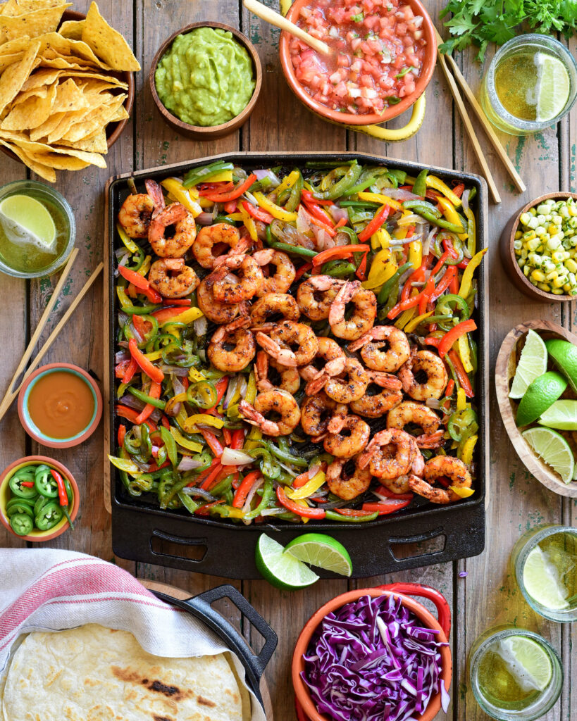 A top down image of a cast iron griddle with grilled tri coloured peppers, onions and shrimp. Surrounding this griddle are the fixings for sizzling steak fajitas. 
