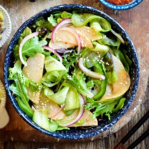 A top down image of a Korean Green Salad with ribbons of cucumbers, slices of Asian pears, onions lettuce, onions and sprinkled with sesame seeds.