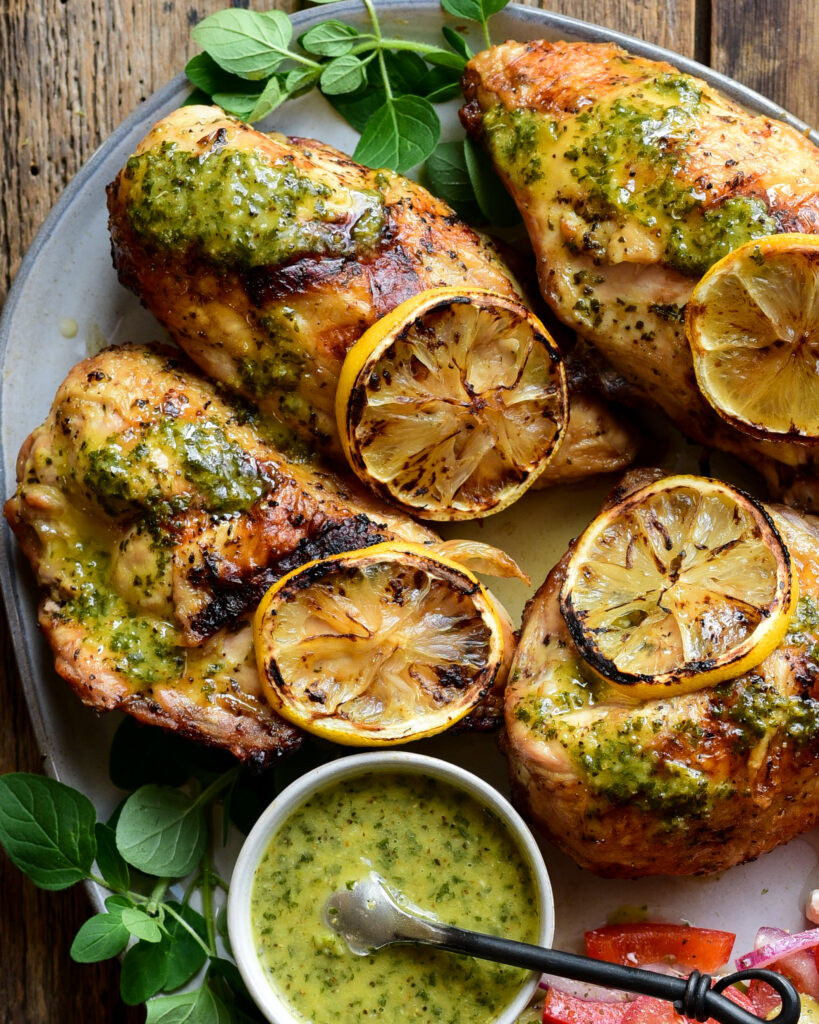 A close up of four grilled lemon-oregano chicken breasts with a bowl of sauce. 