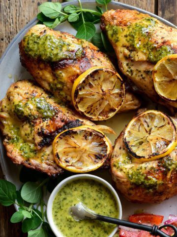A close up of four grilled chicken breasts with a lemon-oregano sauce and grilled lemon slices and a bowl of sauce.