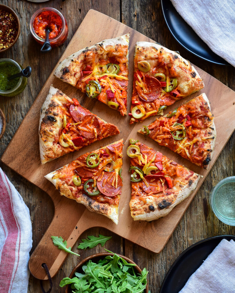 Field Roast Pizza. A top down image of a plant-based pepperoni pizza cut in six slices on a wooded pizza peel. A bowl of arugula and pizza toppings surround the pizza. 