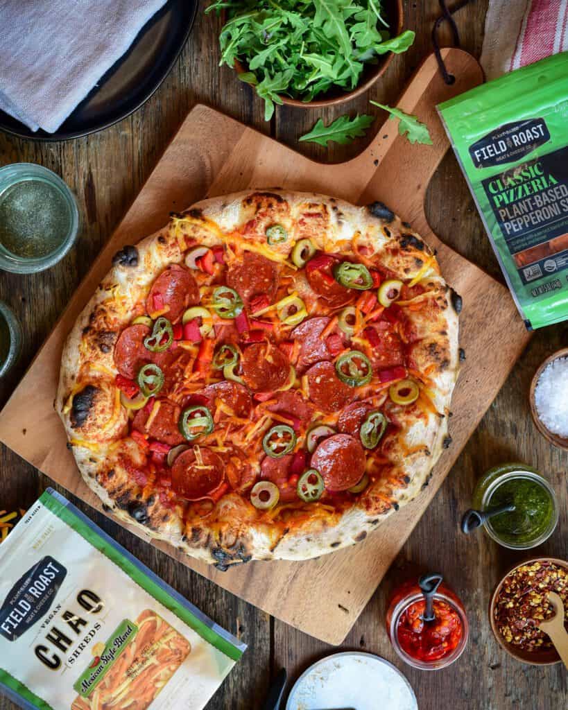 Field Roast Pizza. A top down image of a plant-based pepperoni pizza on a wooded pizza peel. A bowl of arugula, pizza toppings surround the pizza. 