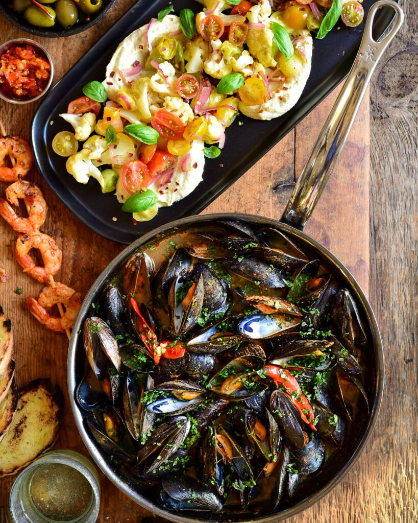 A top down image of Red Thai Curry Mussels in a skillet. A dish of hummus and veggies, shrimp, toast, olives, hot chilli sauce and a glass of wine surround the skillet 