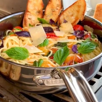 A large pan of cherry tomato spaghetti with garlic toast outside on a grill.