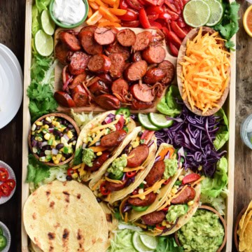 A top down image of a very colourful tray of Bavarian Smokie tacos. Along with the four made up tacos there is a bowl of shredded cheese, a bowl of corn & black bean salsa, a bowl of guacamole, tortillas, sliced grilled sausages, limes, chopped tomatoes and shredded lettuce.