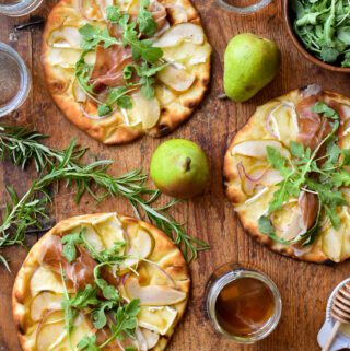 This is a top down image of three Ontario Pear & Prosciutto Naan-Pizzas with a pot of honey, rosemary sprigs, Bartlett pairs and glassed of wine on a wooden cutting board.