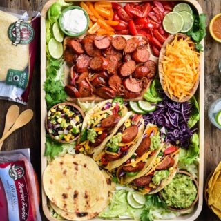 A top down image of a very colourful tray of Bavarian Smokie tacos. Along with the four made up tacos there is a bowl of shredded cheese, a bowl of corn & black bean salsa, a bowl of guacamole, tortillas, sliced grilled sausages, limes, chopped tomatoes and shredded lettuce.