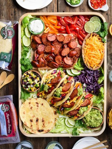 A top down image of a very colourful tray of Bavarian Smokie tacos. Along with the four made up tacos there is a bowl of shredded cheese, a bowl of corn & black bean salsa, a bowl of guacamole, tortillas, sliced grilled sausages, limes, chopped tomatoes and shredded lettuce.