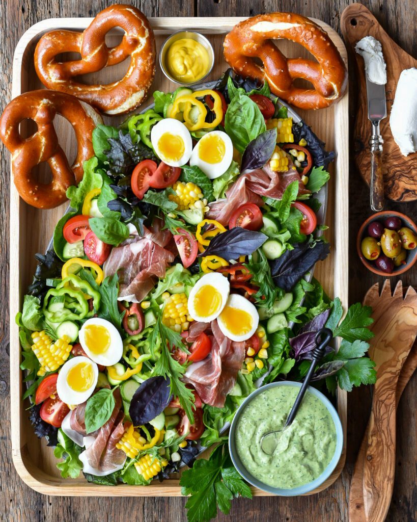 A salad platter filled with soft boiled eggs, prosciutto, greens and pretzels with a bowl of dressing.