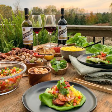 An outdoor setting with ingredients for Spanish lettuce wraps laid out, along with two bottles of wine, wine glasses and plates. A finished lettuce wrap is in the forefront.