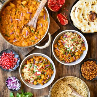 A top down image of Roasted Butternut Squash, Cauliflower and Chickpea Curry. The curry and rice are in separate posts and there are two bowls filled with curry and topped with pomegranate arils, chickpeas, onions and cilantro as well as naan bread.