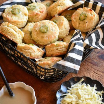 An image of a basket of jalapenos biscuits nestled in a black and white striped tea towel. There is a dish of gravy and a plate of shredded cheese next to this basket.