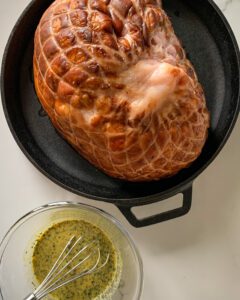 An image of a ham in a round cast iron pan with a bowl sauce and a whisk.
