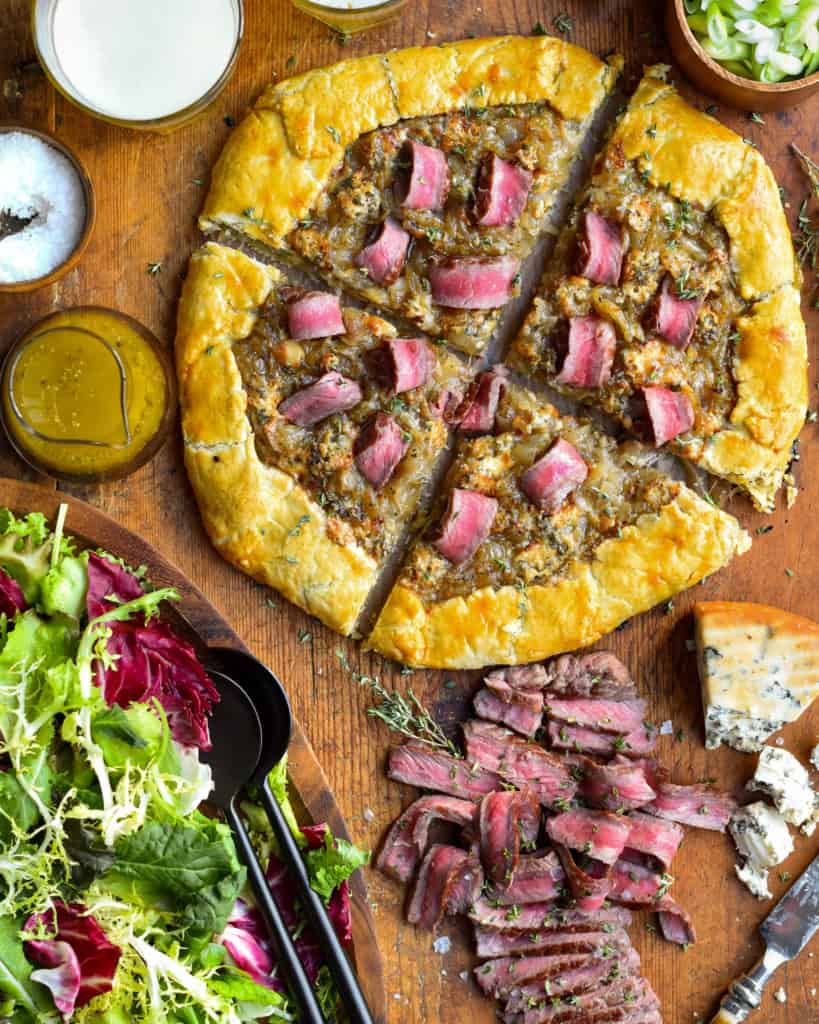 An onion and steak galette sliced into four pieces and served with a salad and extra steak slices.