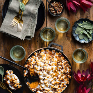 Sweet Potato Casserole with toasted marshmallows in a cast iron pan. A scoop is take out to reveal the inside and a portion is plated. Glassed, dishes, toppings and flowers surround the dish.
