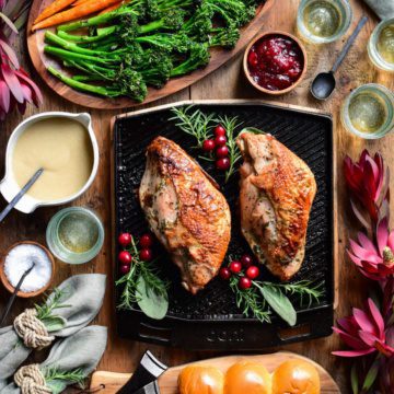 A top down image of two cooked turkey breasts on a skillet with herbs and cranberries. Broccolini, carrots, gravy, buns, cranberry sauce, wine glasses and florals surround this skillet.