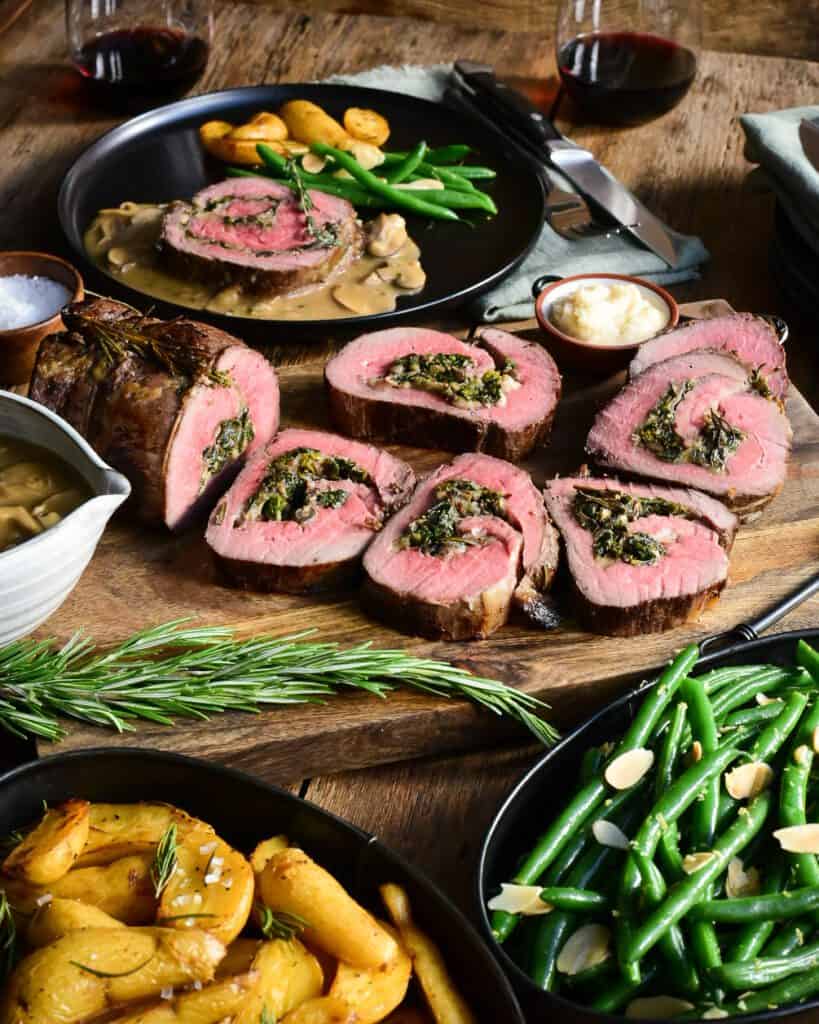 A sliced stuffed roast beef surrounded with a bowl of gravy, a dish of green beans, potatoes, horseradish, salt, napkins and cutlery