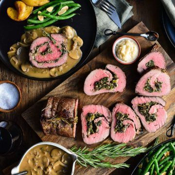 A sliced stuffed roast beef surrounded with a bowl of gravy, a dish of green beans, potatoes, horseradish, salt, napkins and cutlery