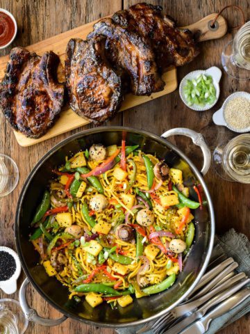 A top down image of a wok with a brightly coloured stir fry beside a cutting board with four beautifully charred pork chops.