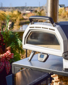 The Ooni Karu 16 pizza oven preheating on a beautiful day with a festive planter, trees and pond in the background.