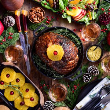 An image of a baked ham in a cast iron skillet with a pineapple on top. There is also winter salad, more baked pineapples a carving knife set and holiday sprigs on the table.