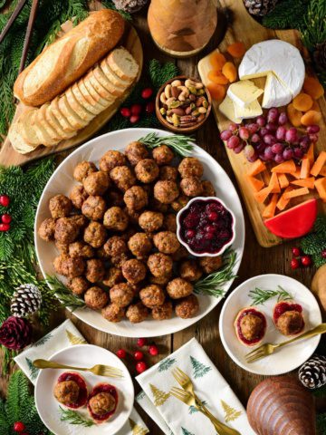 A top down image of a round platter filled with meatballs and bowl of cranberry sauce. A sliced baguette, a cheese board, plates and festive decoration surround this platter.
