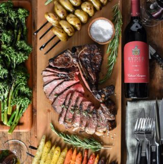A top down image of a sliced t-bone steak, skewered potatoes and carrots, broccolini and a bottle of red wine and glasses.