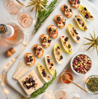 A marble tray of seven cheese crostinis, and endive boats with a block of cheese all topped with walnuts, honey, dried cherries and rosemary. Surrounded by glasses of pink Champaign, rosemary, honey and twinkle lights.