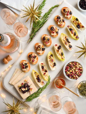 A marble tray of seven cheese crostinis, and endive boats with a block of cheese all topped with walnuts, honey, dried cherries and rosemary. Surrounded by glasses of pink Champaign, rosemary, honey and twinkle lights.