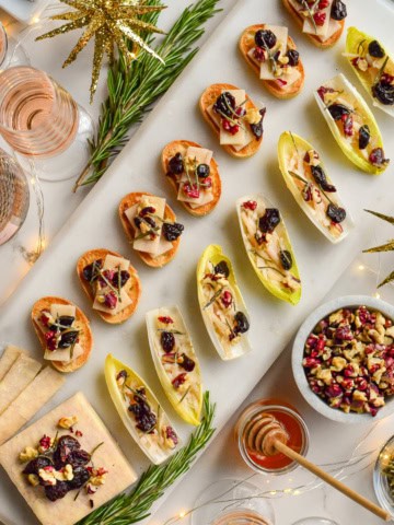 A marble tray of seven cheese crostinis, and endive boats with a block of cheese all topped with walnuts, honey, dried cherries and rosemary. Surrounded by glasses of pink Champagne, rosemary, honey and twinkle lights.