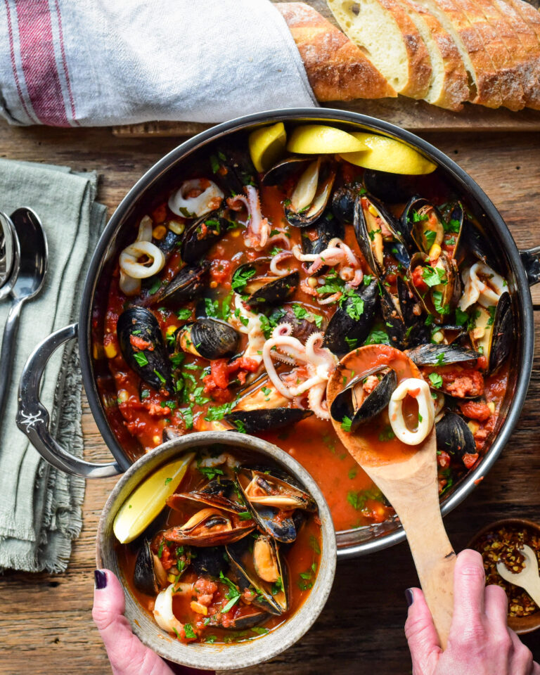 A top down image of large pot of mussels and squid in a tomato broth. A portion is being spooned out into a bowl.