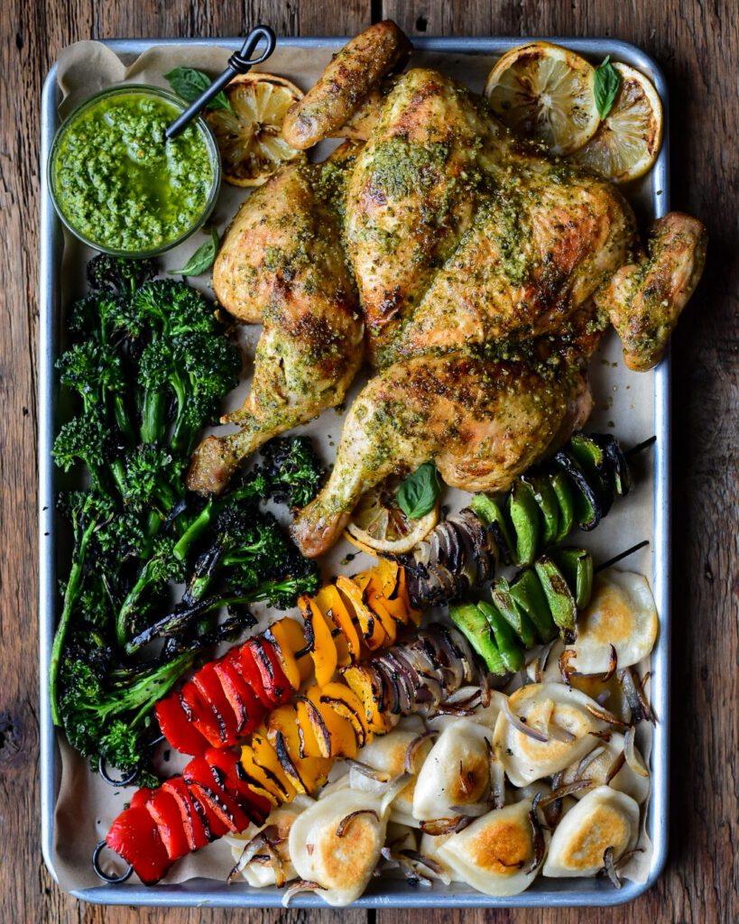 A sheet pan with a grilled pesto spatchcocked chicken, charred broccolini, grilled peppers & onions and fried pierogi with onions.