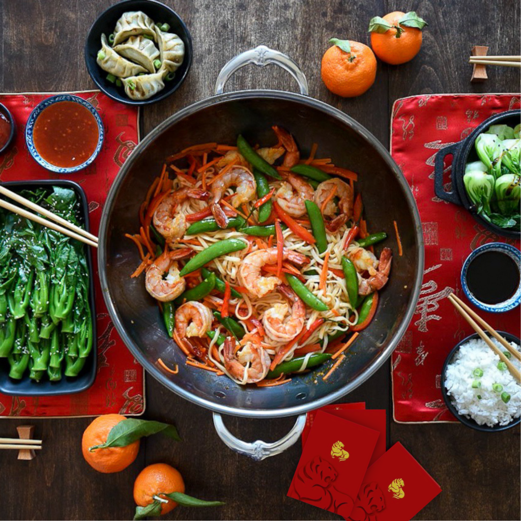 A top down image of a wok with a shrimp and noodle stirfry. An Asian themed table setting surround the wok.