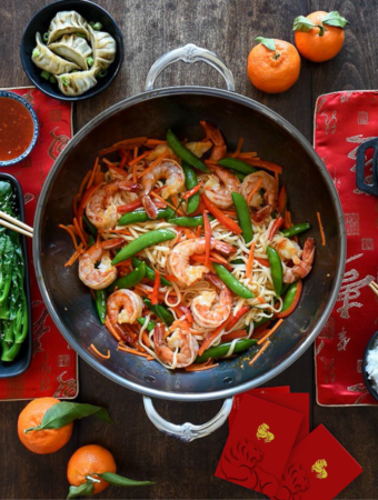 A top down image of a wok with a shrimp and noodle stirfry. An Asian themed table setting surround the wok.