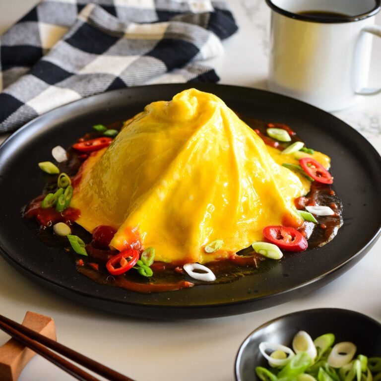 A domed and swirled tornado omelette (Omurice) over a pile of rice (not shown) with sauce and toppings on the base.