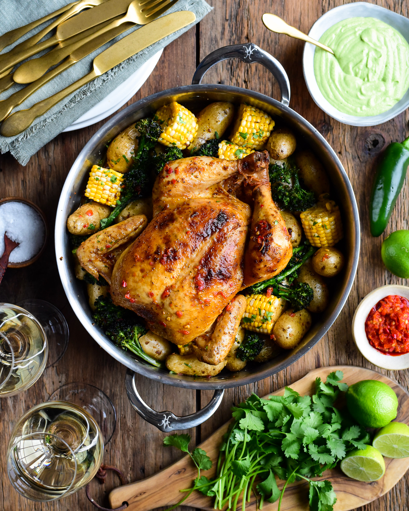 Top down image of a roasted chicken in a pan with roasted vegetables and bowl of green sauce.