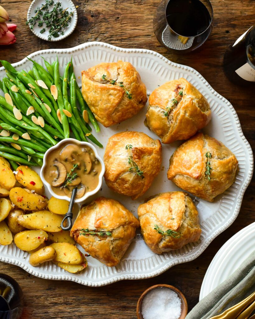 An oval platter with six individual ground beef wellingtons, a bowl of marsala mushroom sauce, potatoes and green beans.
