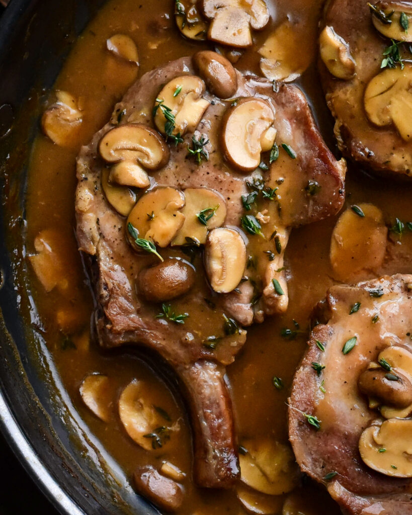 A close up of a pork chop in a pan filled with a creamy mushroom sauce.