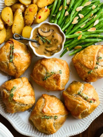 An oval platter with six individual ground beef wellingtons, a bowl of marsala mushroom sauce, potatoes and green beans.