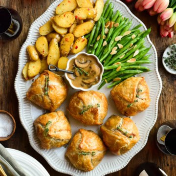 An oval platter with six individual ground beef wellingtons, a bowl of marsala mushroom sauce, potatoes and green beans.