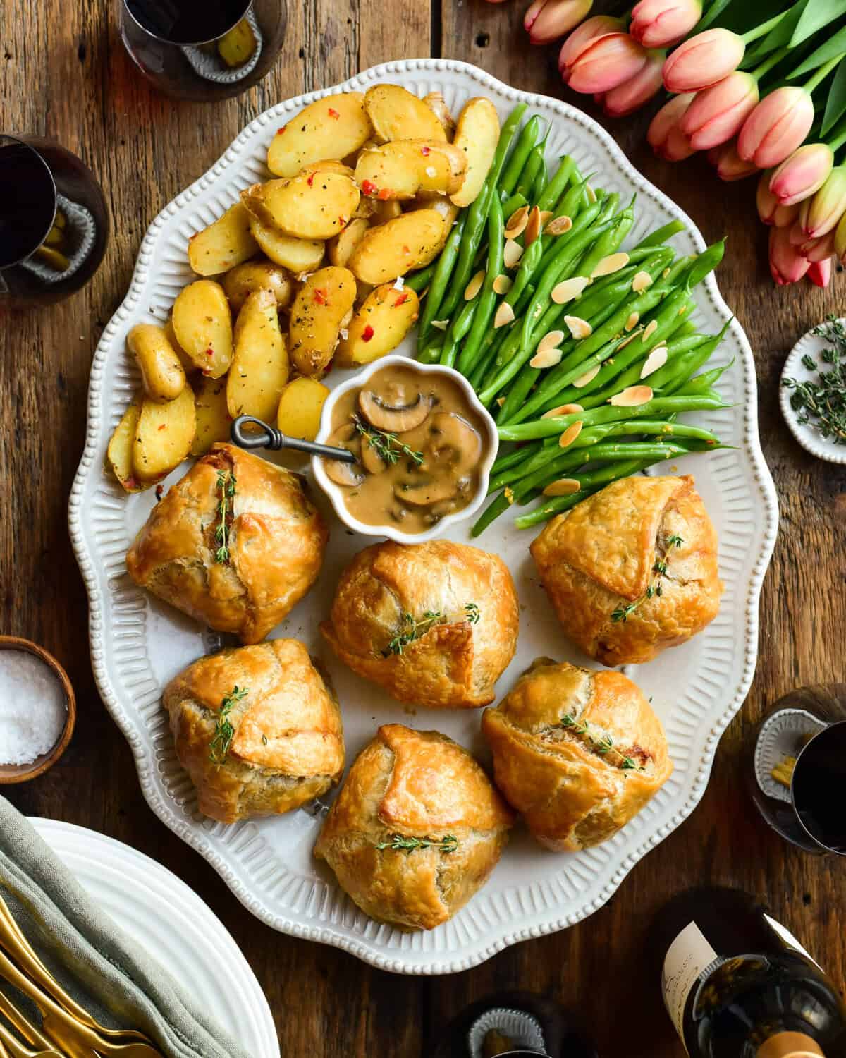 An oval platter with six individual ground beef wellingtons, a bowl of marsala mushroom sauce, potatoes and green beans.