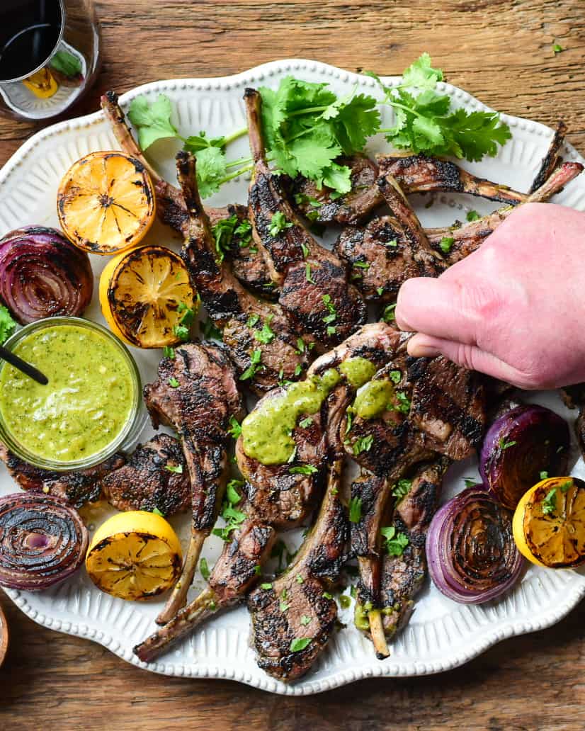 A platter of lamb chops with a green sauce with someone holding a sauced chop. 